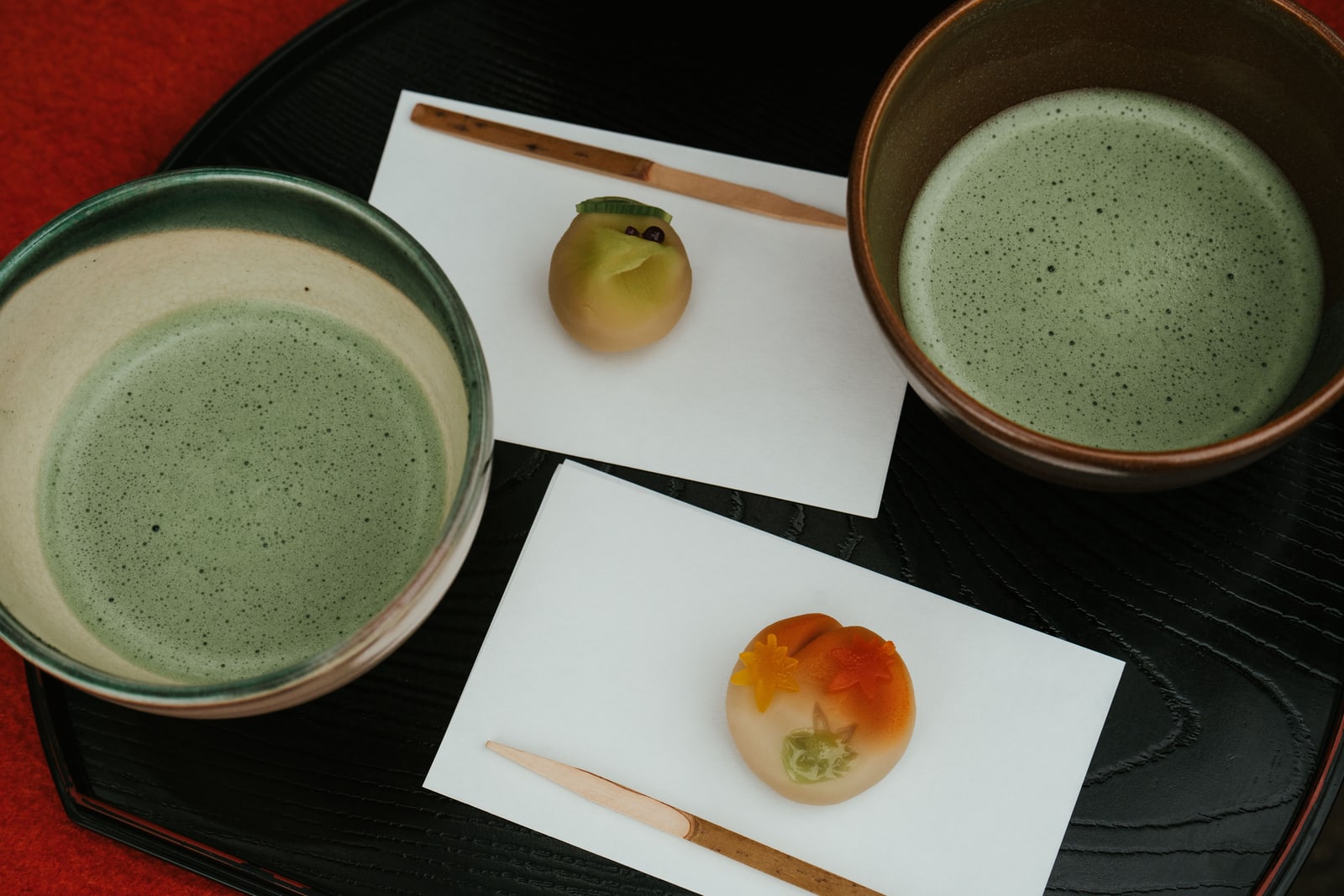 Two bowls of matcha and two wagashi sweets on individual papers, arranged on a black lacquer tray.