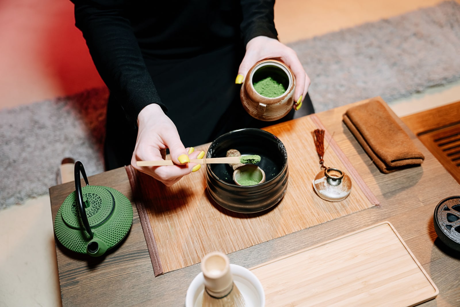 Hands scooping matcha powder from a caddy into a bowl beside a whisk, an iron kettle and a cloth on a wooden table.