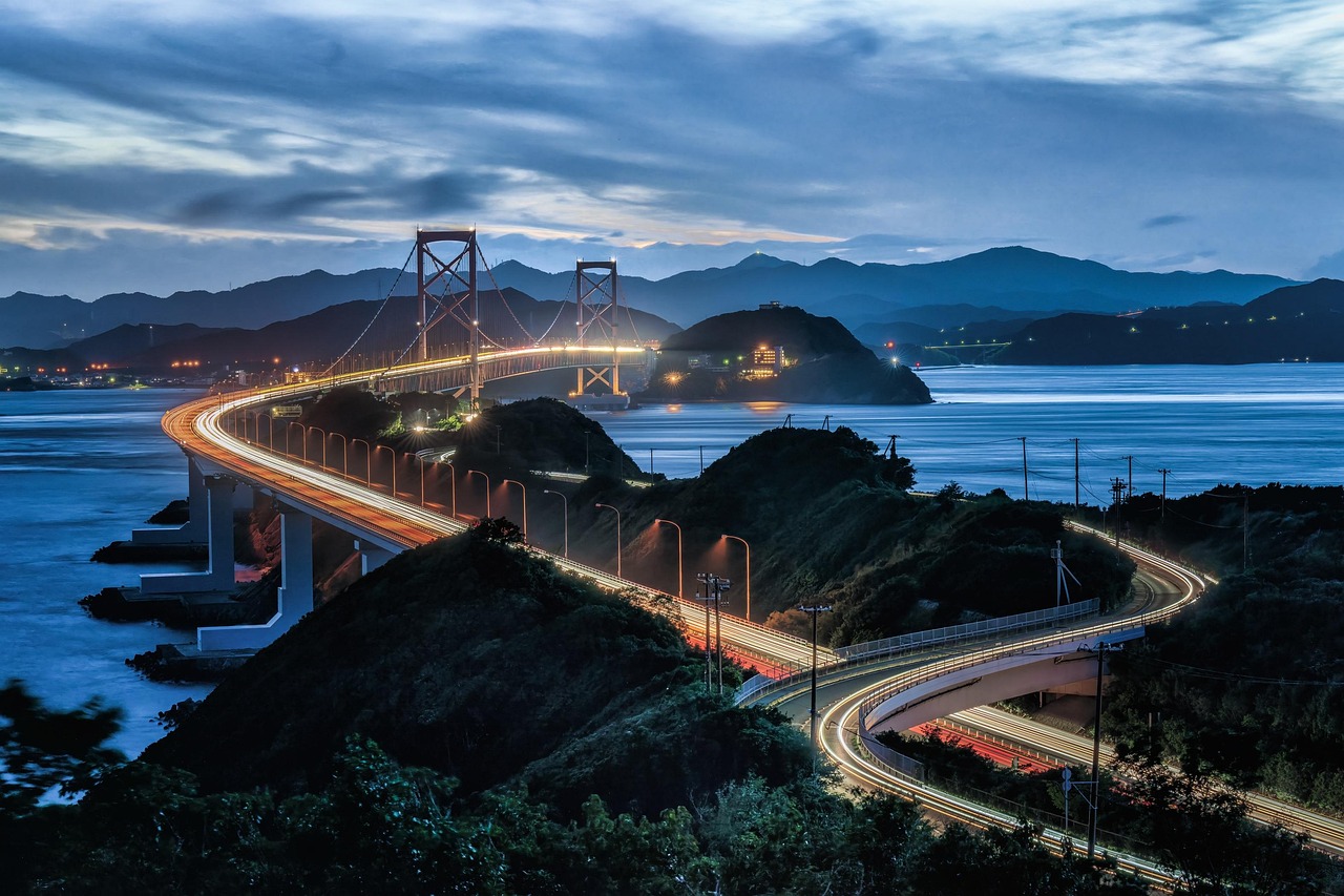 A suspension bridge over a strait at dusk, with car light trails on the curving approach road