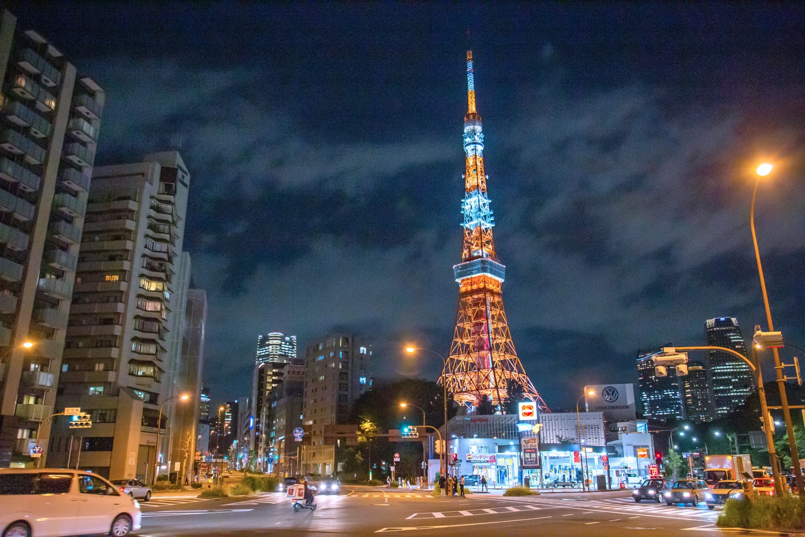 Tokyo Tower lit at night above a street intersection in Minato, with traffic and shop lights in the foreground.