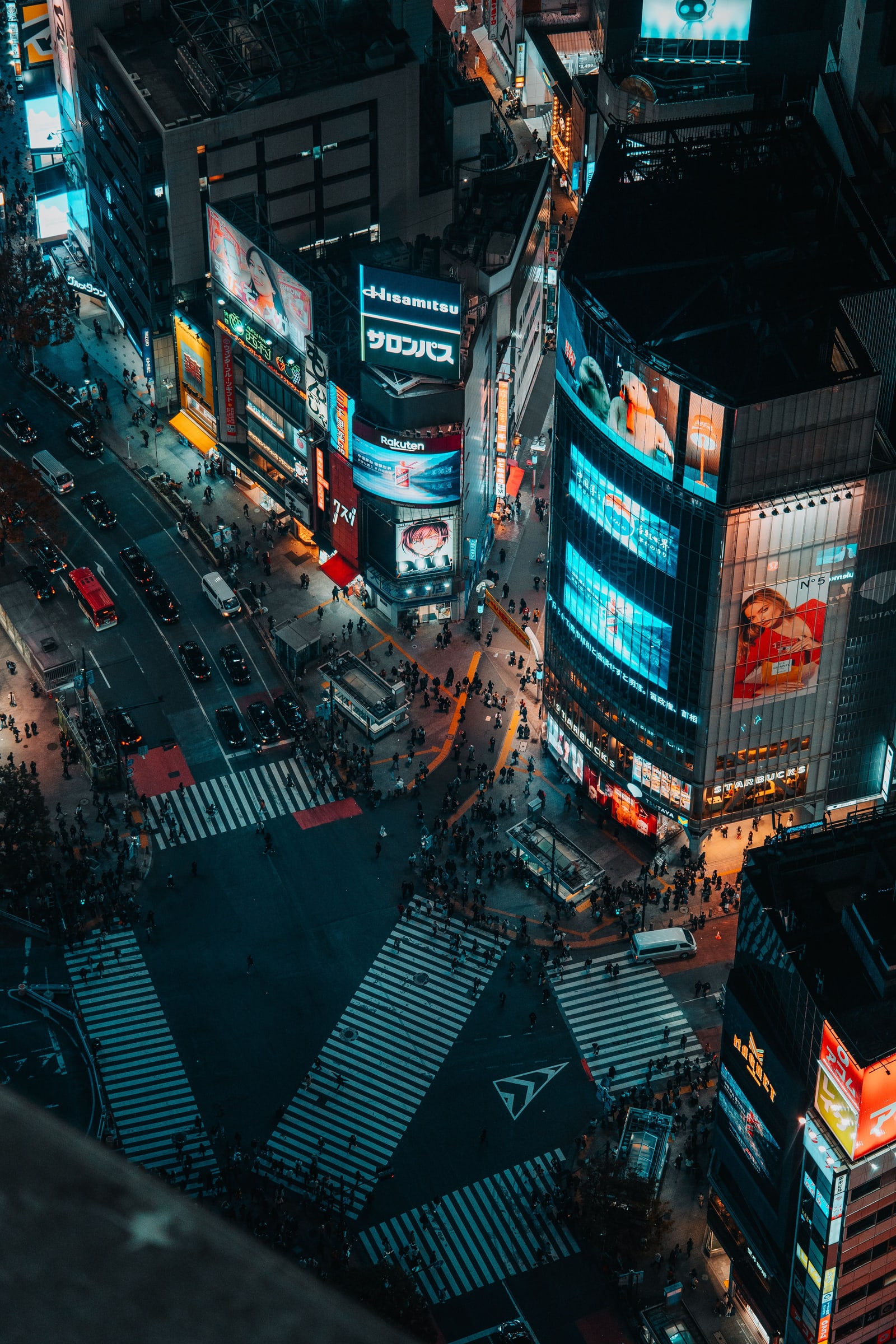 Shibuya Crossing at night seen from far above, the striped crosswalks and lit billboards below.