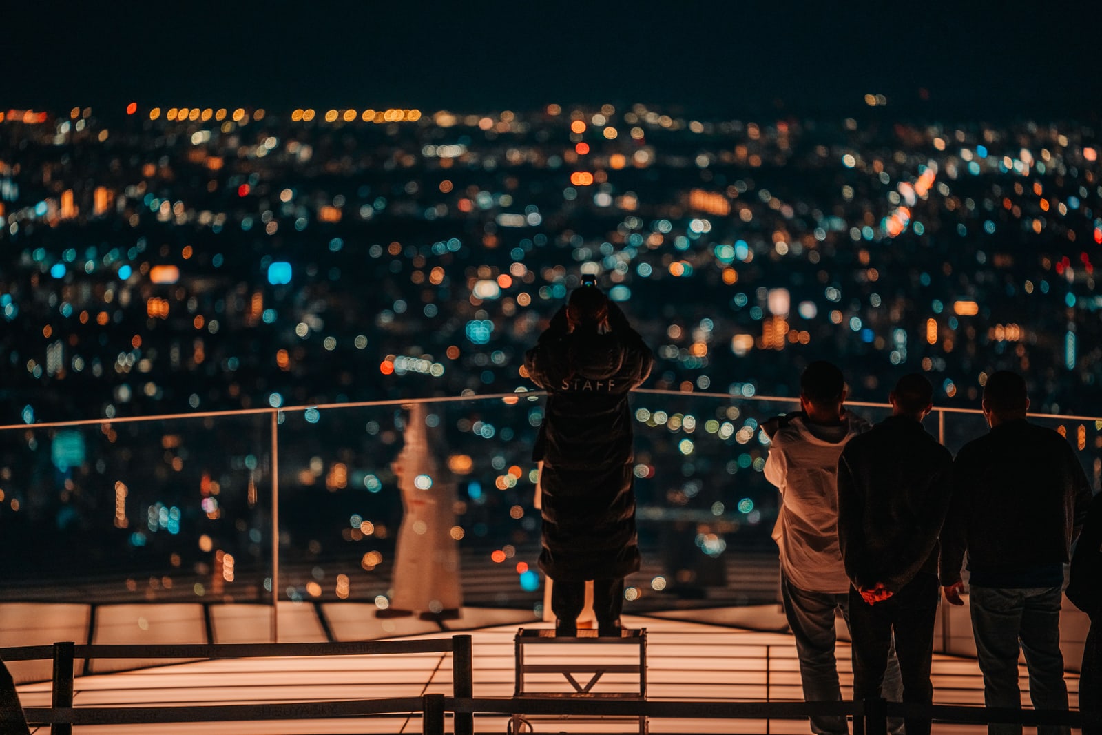 People at the railing of an open-air rooftop deck at night, one holding a phone up to the city lights below.