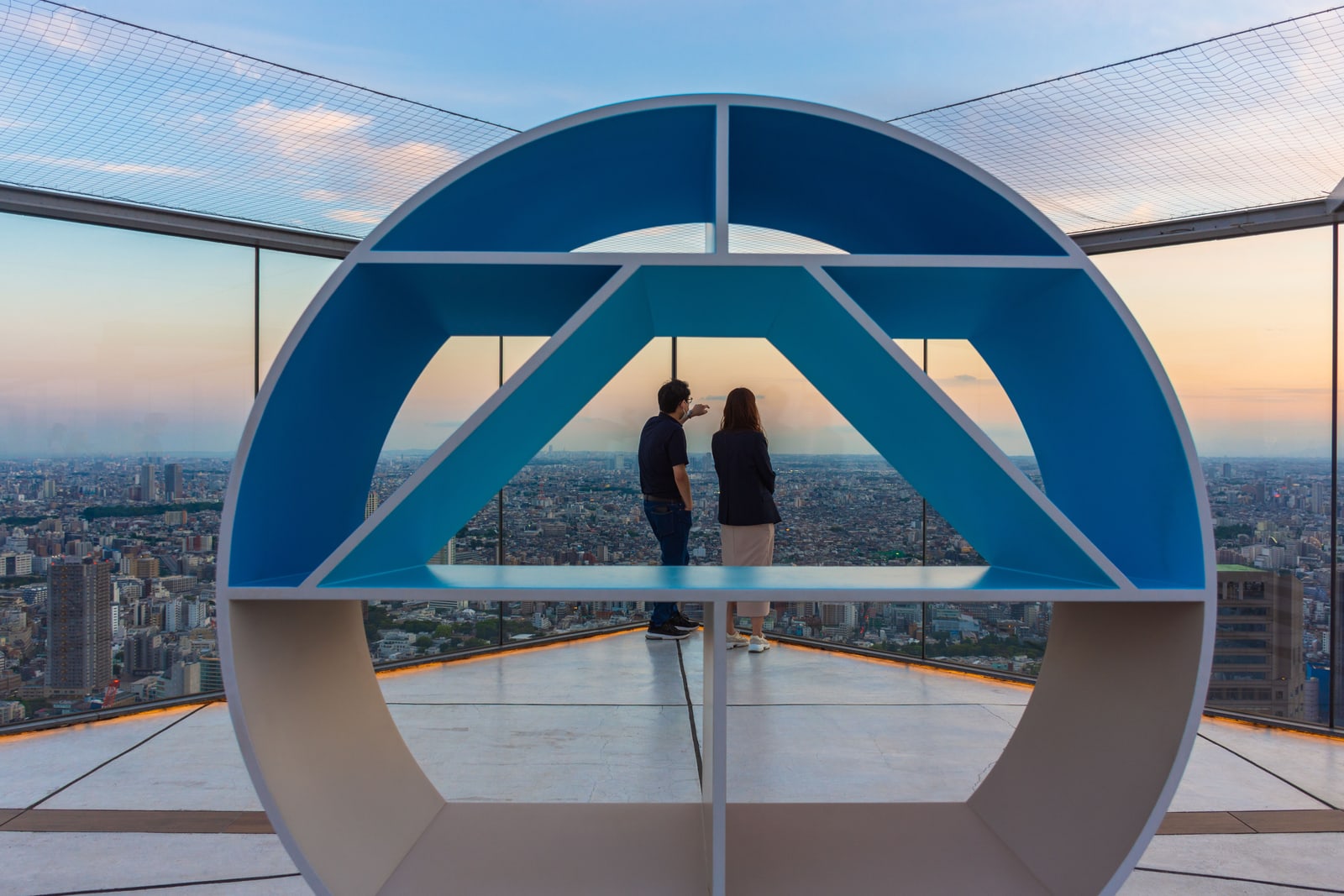 Two people looking out from inside a round blue installation on an open-air rooftop deck at sunset.