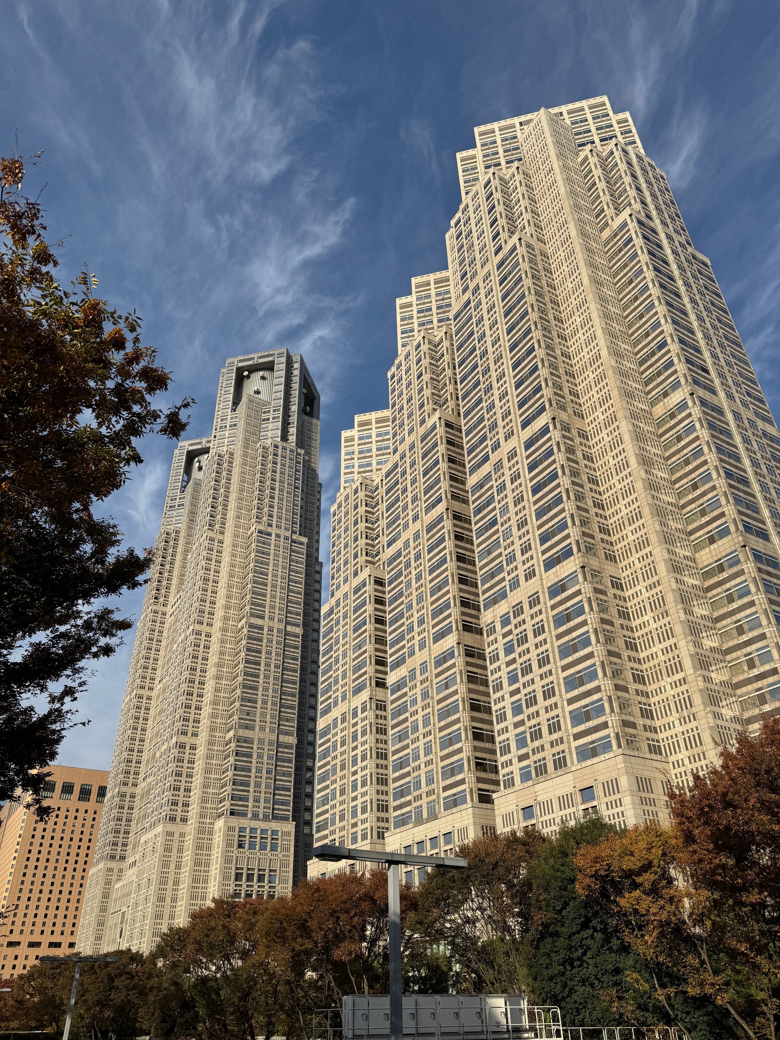 The twin towers of the Tokyo Metropolitan Government Building seen from below against a blue sky, autumn trees in the foreground.