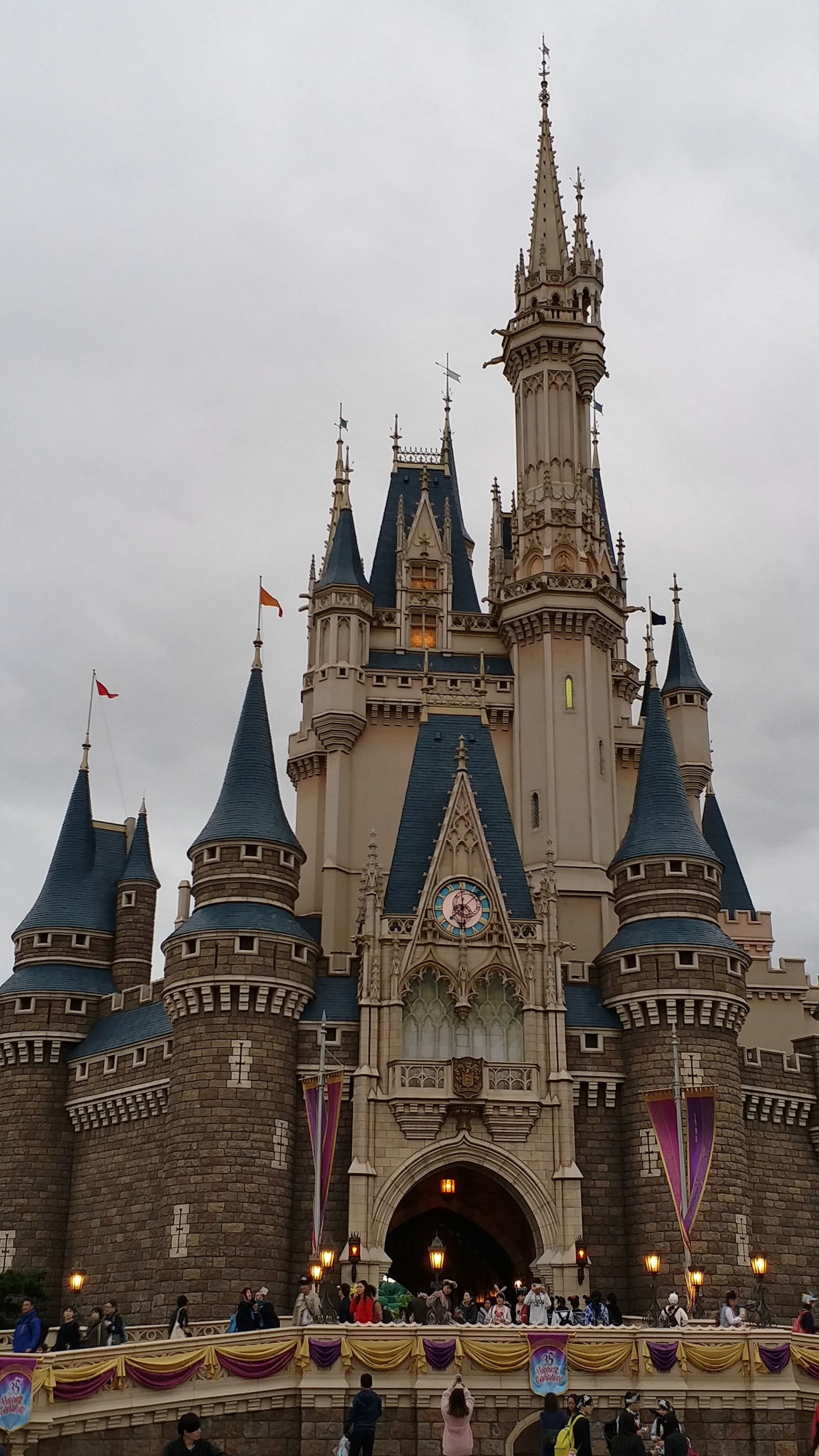 A low-angle view of Cinderella Castle at Tokyo Disneyland under an overcast sky, visitors on the entry bridge below.