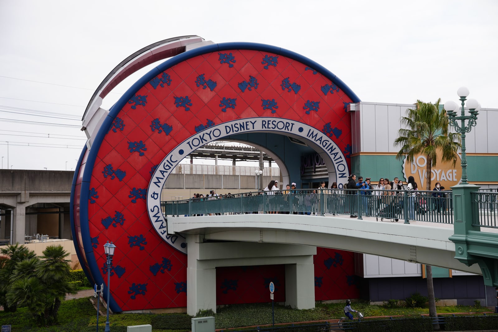 The Tokyo Disney Resort entrance building with the Bon Voyage store, TOKYO DISNEY RESORT lettering on the arch, and visitors crossing the pedestrian bridge.