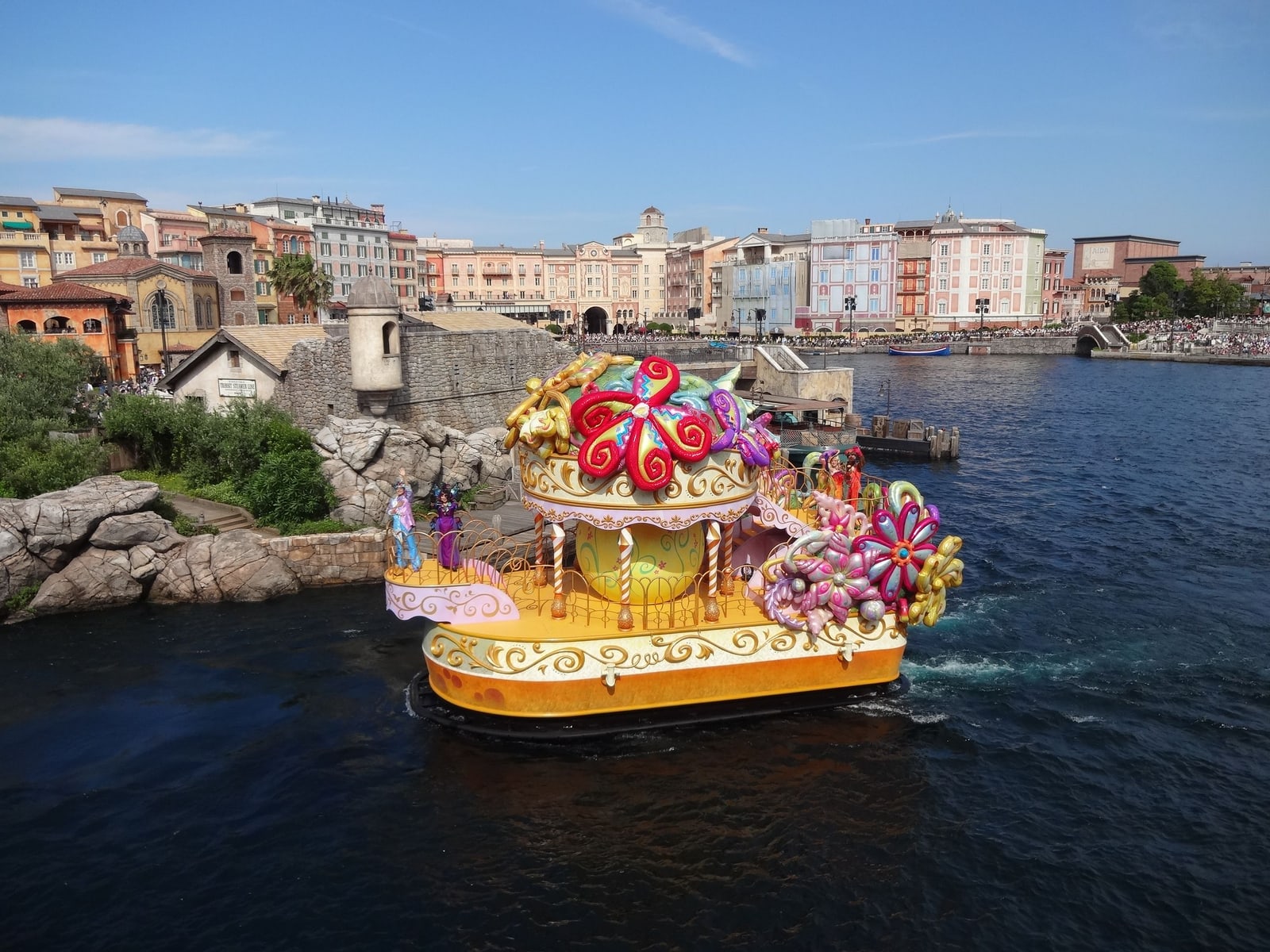 A colorful parade float on the water of Tokyo DisneySea's Mediterranean Harbor, Italian-style buildings behind.