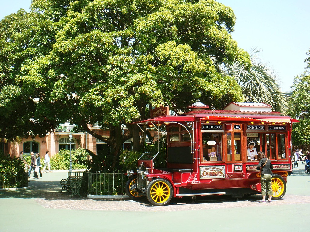 A vintage-style red churro cart under a large tree at Tokyo Disneyland, with a vendor and a customer beside it