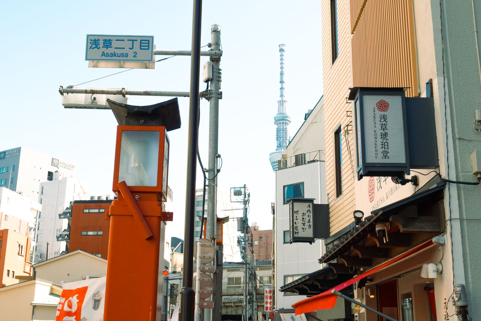 A low-angle Asakusa street with Tokyo Skytree in the distance, overhead cabling, and a municipal street plate reading 浅草二丁目 / Asakusa 2.