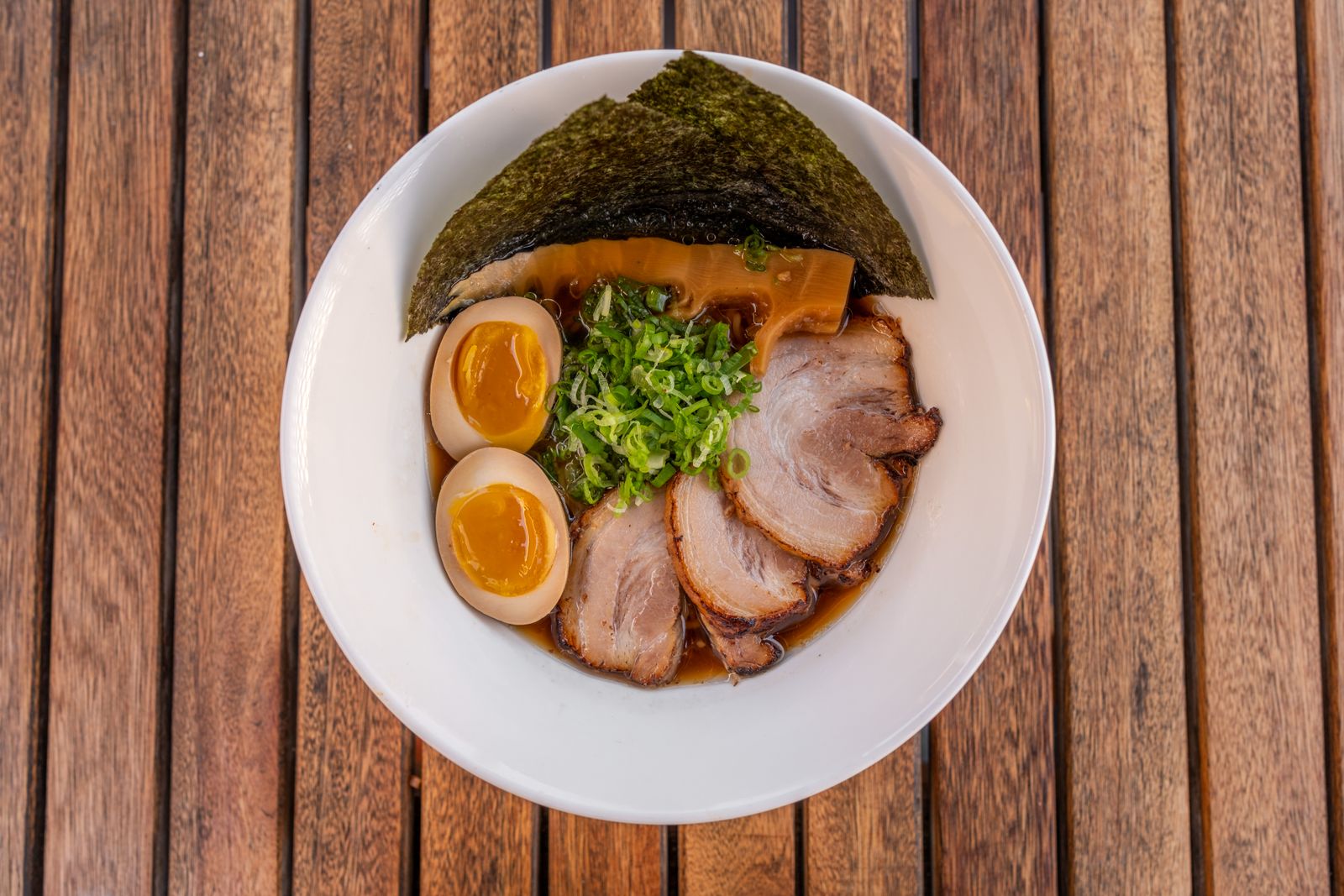 A bowl of shoyu ramen seen from above on a slatted wooden table: clear brown broth, chashu, halved marinated egg, menma, nori and scallion.