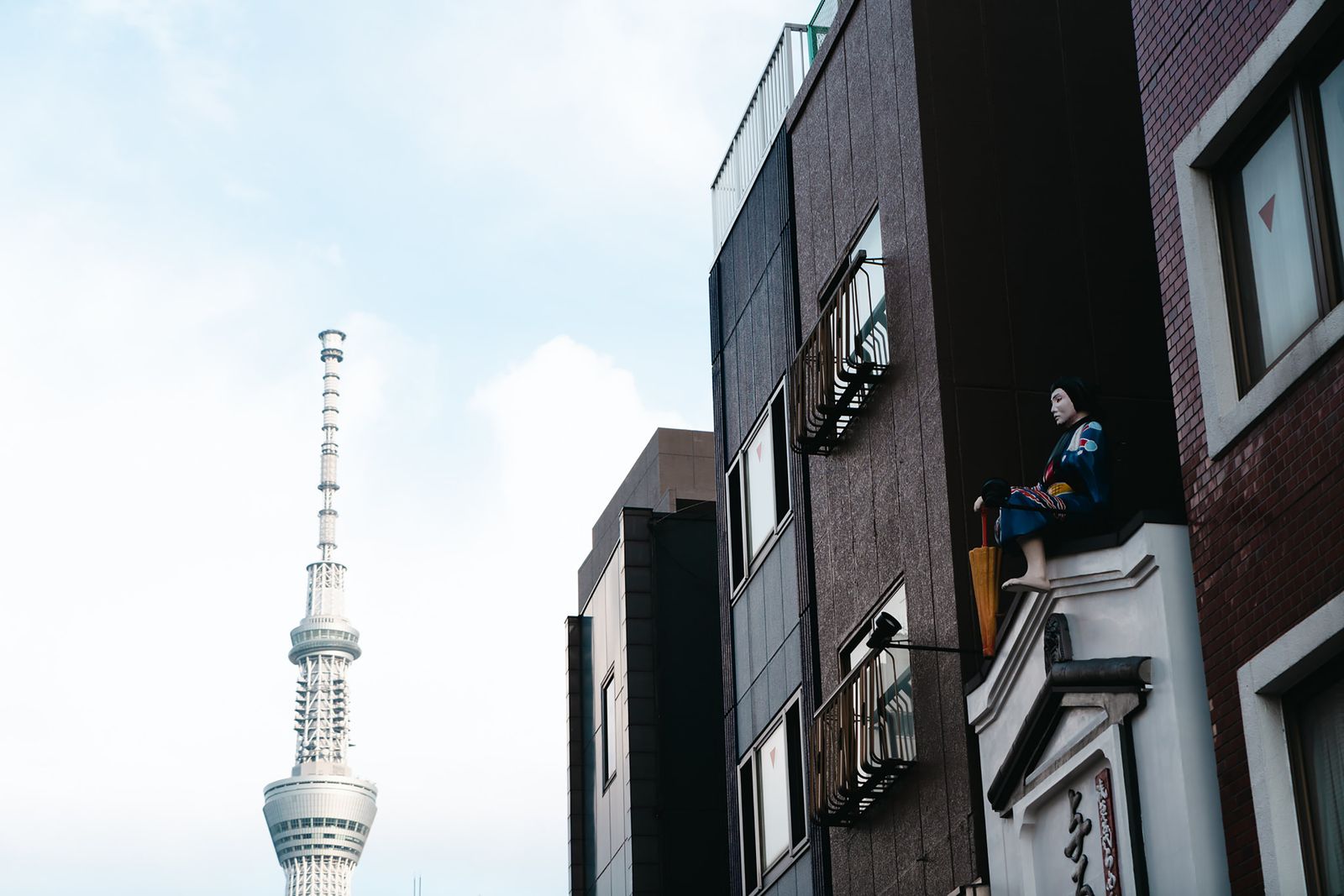 A low upward view of Tokyo Skytree at the left against open sky, ordinary mid-rise blocks at the right, and a painted seated figure in a blue kimono with an umbrella on a ledge.
