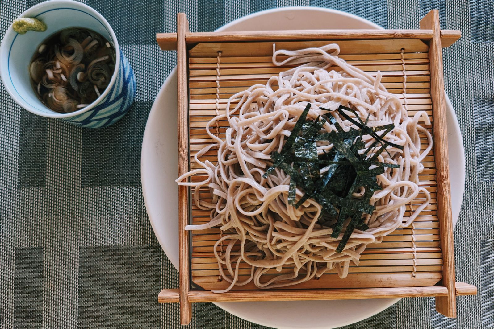 Zaru soba seen from above: buckwheat noodles on a slatted bamboo tray with shredded nori, beside a cup of dipping broth with sliced negi and a dab of wasabi.