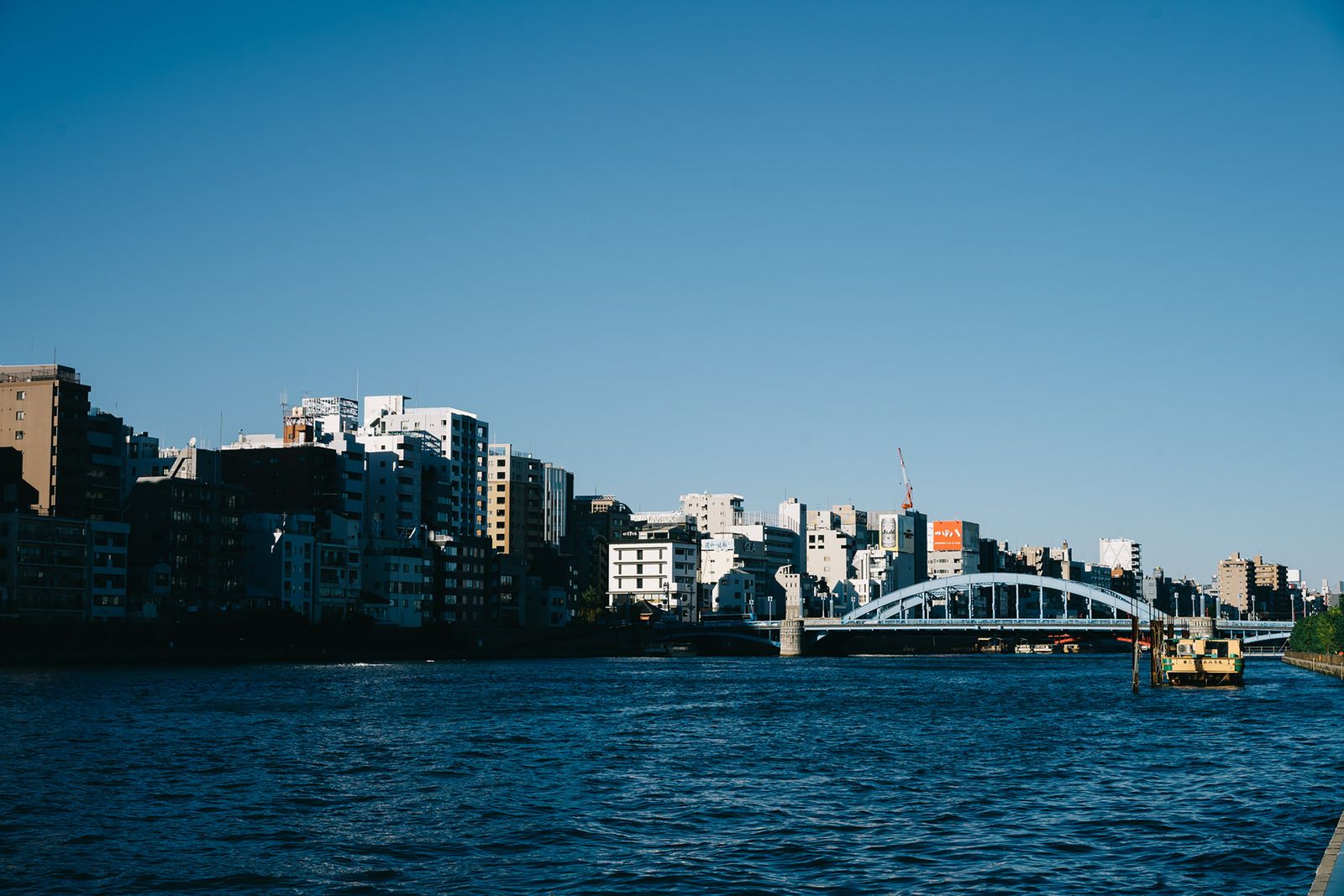A broad river in Tokyo with apartment and office blocks along the left bank, a pale steel arch bridge at the right and a small moored boat.