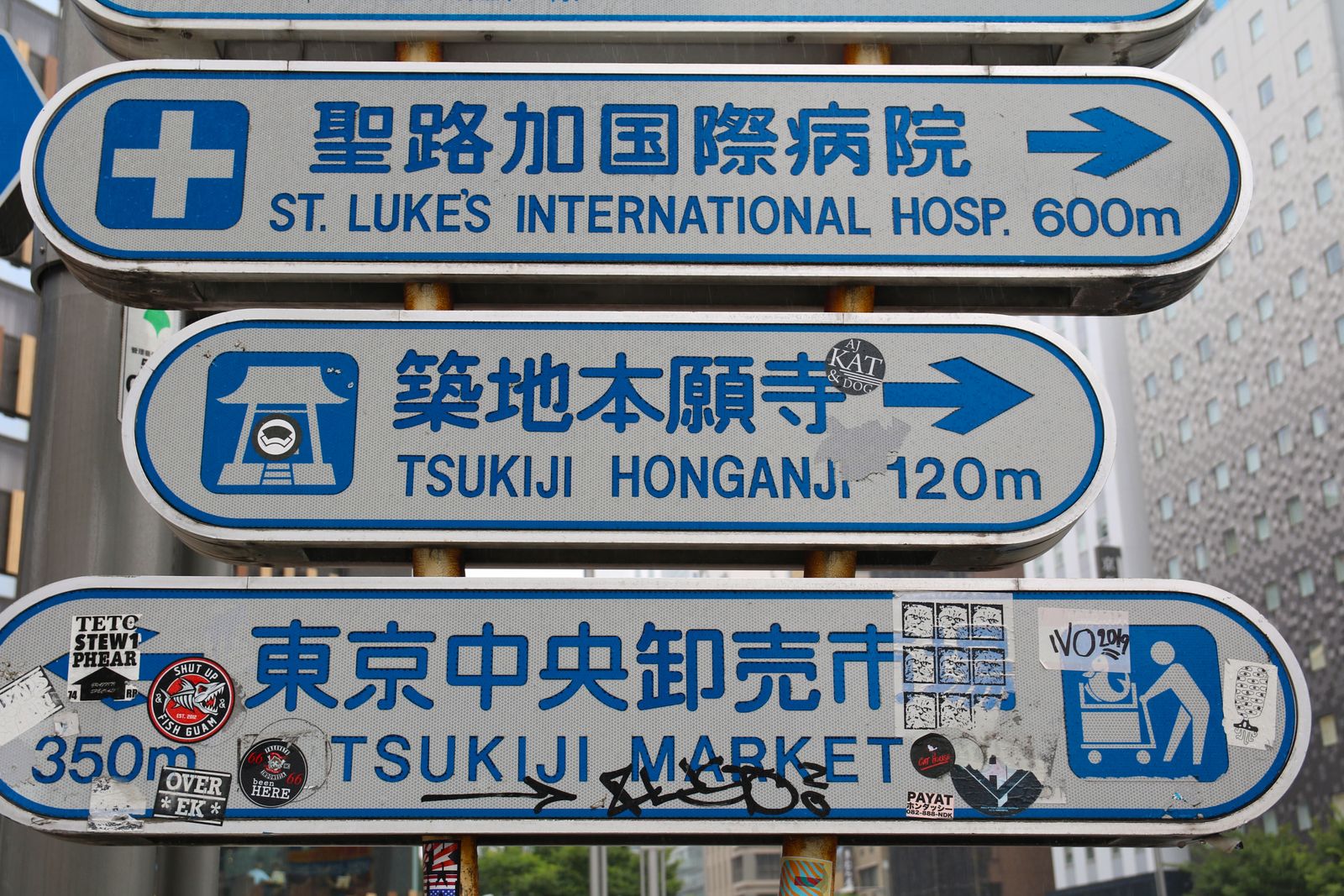A stack of city direction signs on a building wall pointing to St. Luke's International Hospital, Tsukiji Hongwanji and Tsukiji Market.