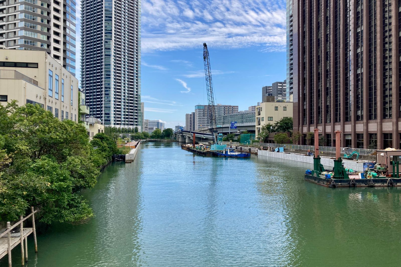 A still green canal running away from the camera between residential towers, trees over the left bank, a crane and a work barge against the right bank.