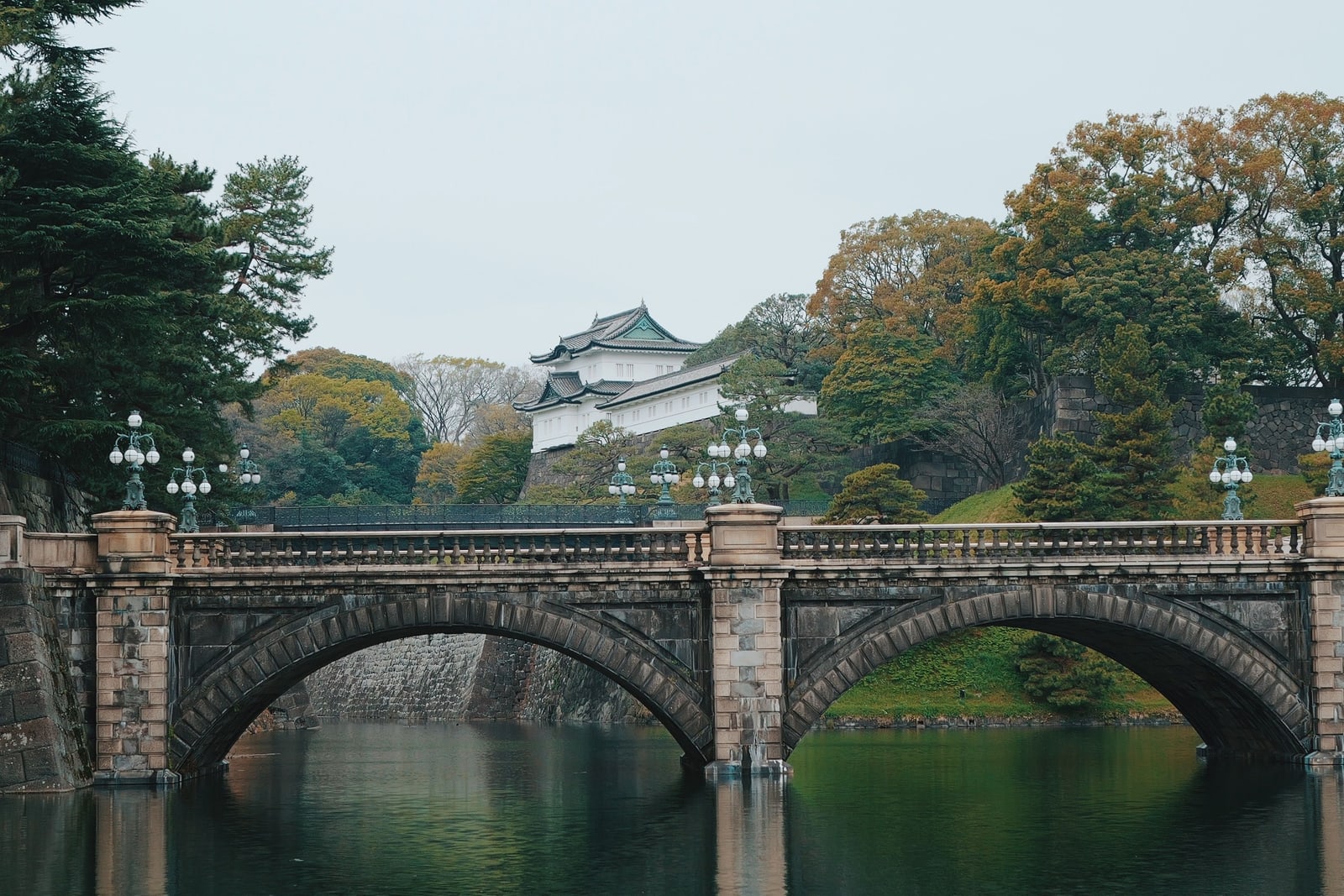 A two-arch stone bridge with lamp posts over the Imperial Palace moat, a white turret and stone wall above it, pines and autumn-coloured trees under an overcast sky