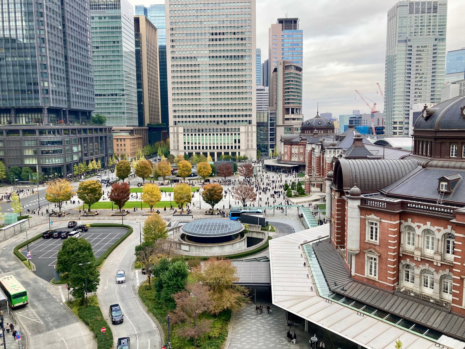 The plaza and red-brick Marunouchi building of Tokyo Station seen from above in autumn, with trees in yellow and red and office towers all round