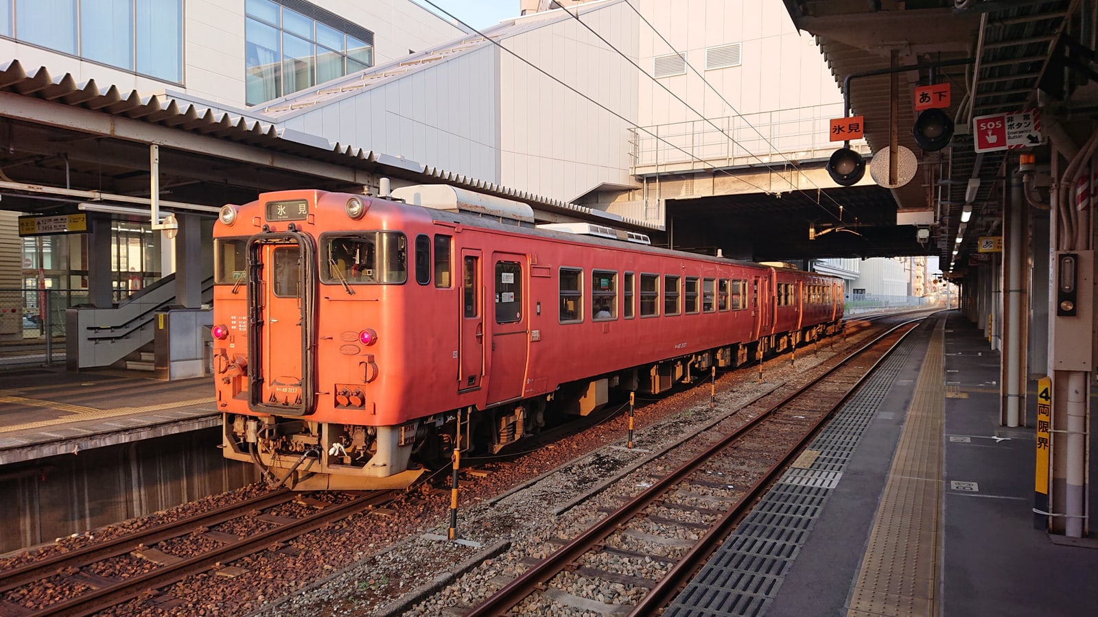 An orange-red local diesel train bound for Himi at a station platform in Toyama Prefecture, under a long canopy in evening light