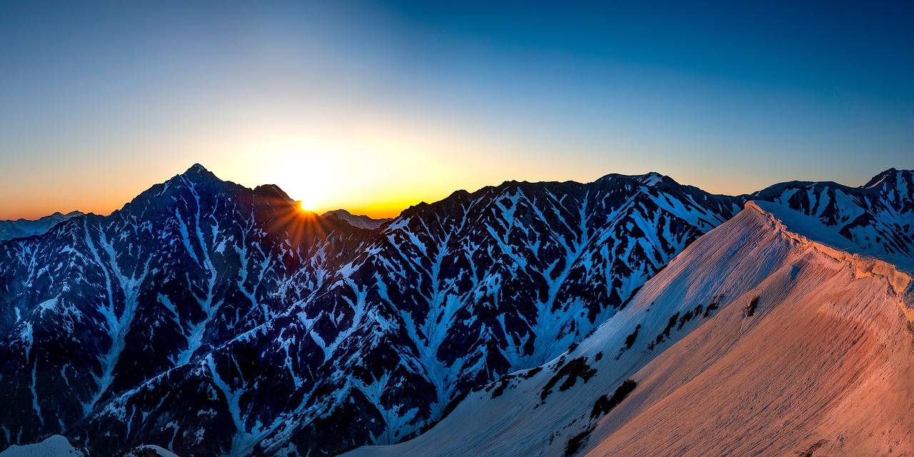 Snow-streaked peaks of the Northern Alps at sunrise, the sun on the skyline and a pink-lit snow ridge in the foreground