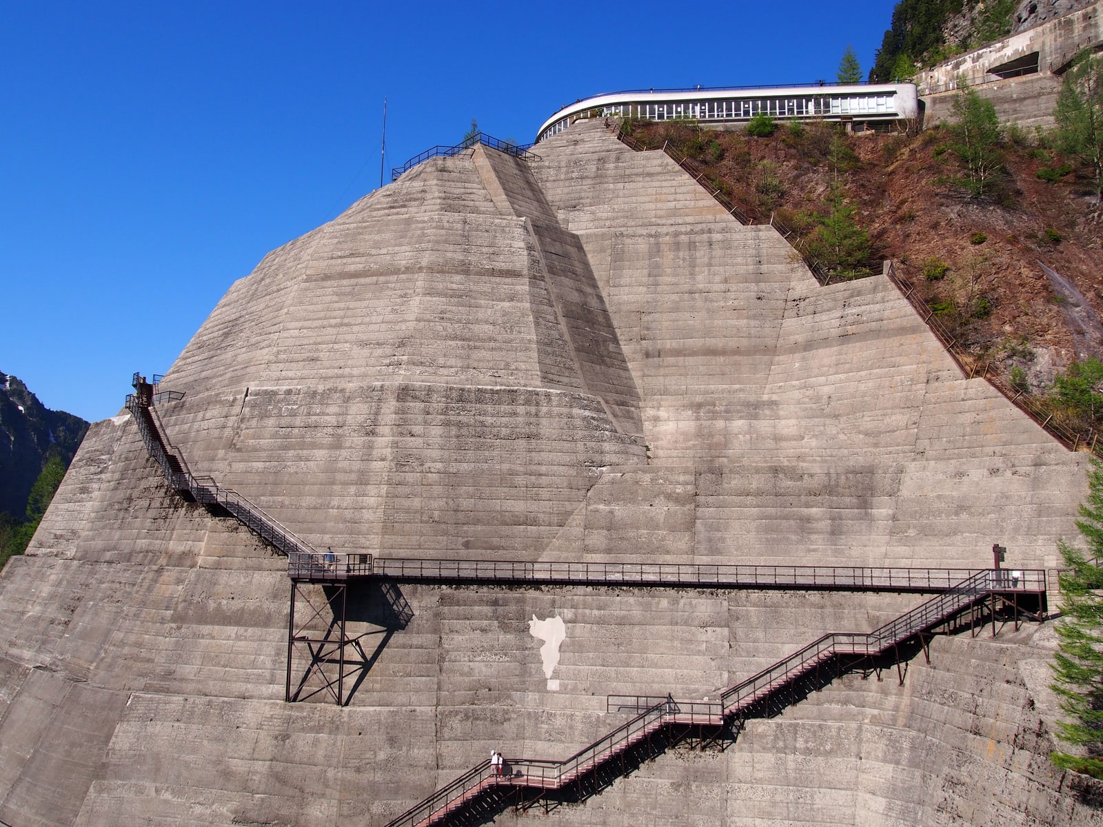 The stepped grey concrete wall of Kurobe Dam with zig-zag steel staircases and a walkway across it, deep blue sky
