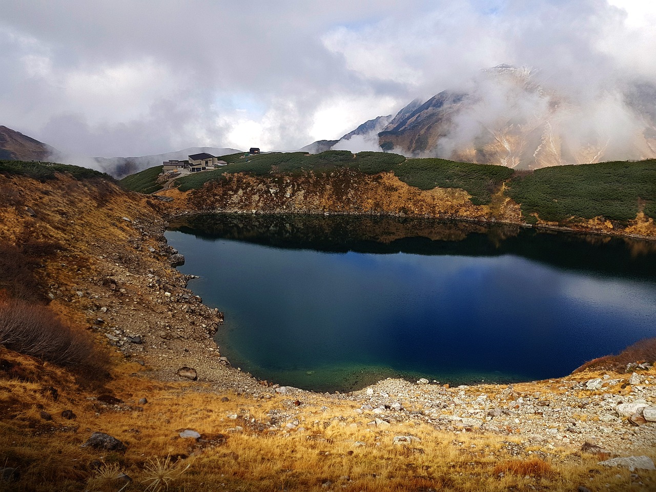 Mikurigaike pond on the Murodo plateau, deep blue in a rocky bowl with golden autumn grass, a lodge on the ridge and mist over the mountains