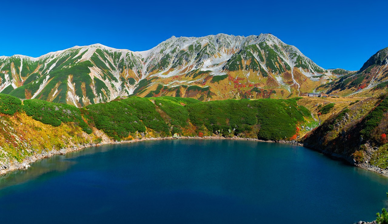 A dark-blue mountain lake below a snow-dusted ridge in the Tateyama area, green creeping pine and red autumn leaves on the slopes, deep blue sky