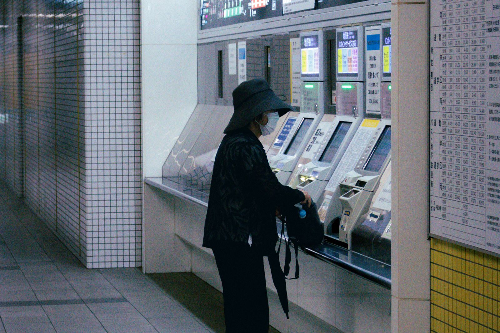 A single figure in a wide-brimmed hat and mask standing at a row of subway ticket machines in Kyoto, a printed fare table on the wall beside them.