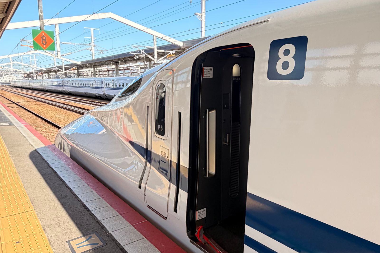 The nose of a white N700-series Tokaido Shinkansen, car 8, standing at a Kyoto Station platform under a clear sky, with a second train on the far track.