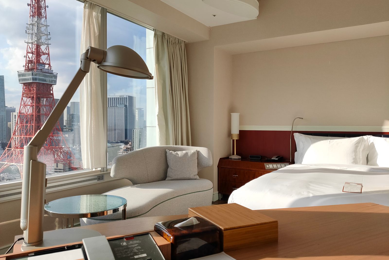 A hotel room with a made double bed and a desk lamp, Tokyo Tower filling the window.
