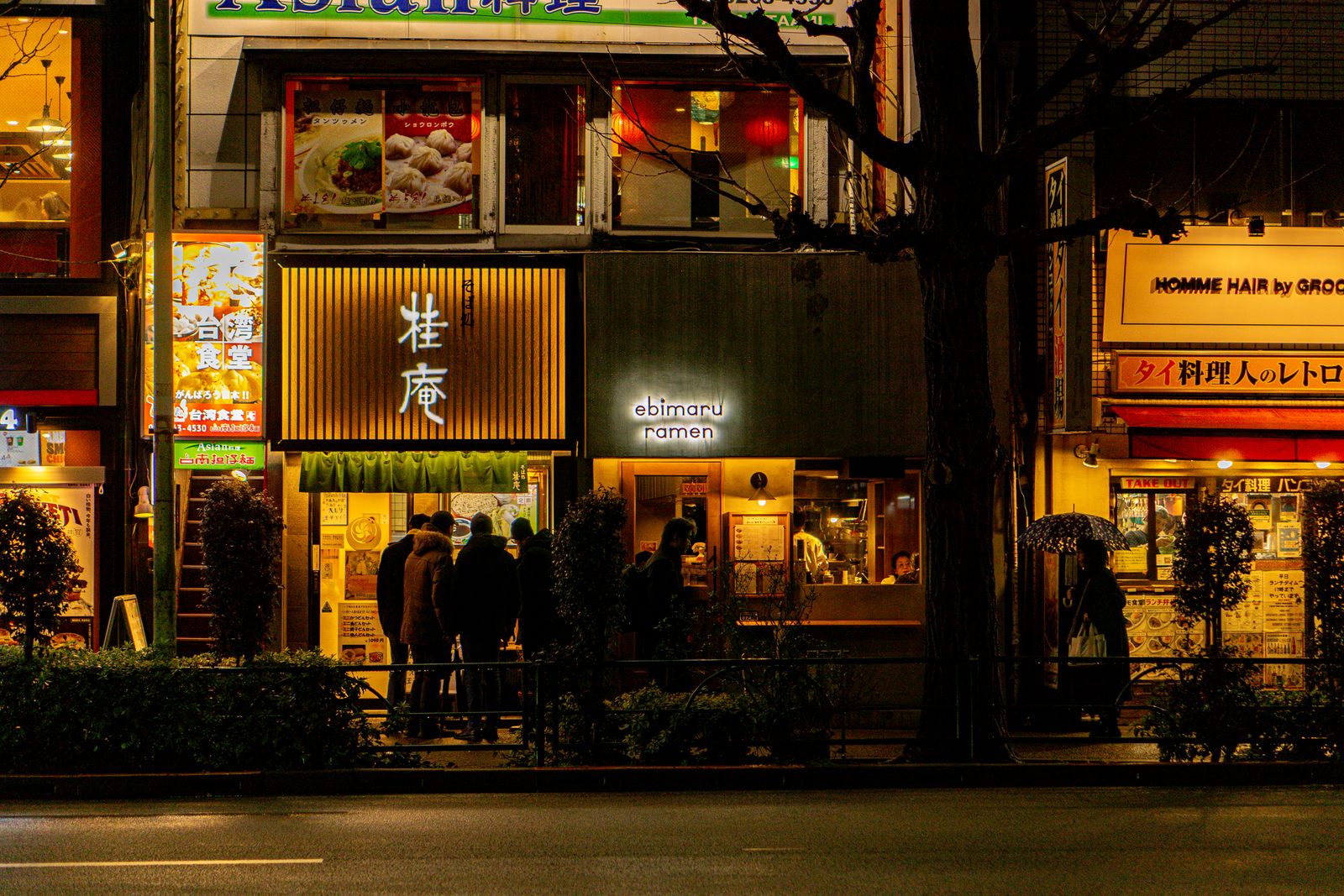 A row of small restaurants lit at night on a Tokyo street, a short queue outside