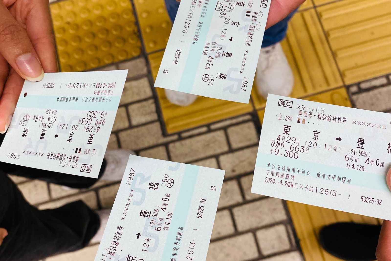 Four hands seen from above, each holding its own Shinkansen ticket, over the yellow tactile paving of a station floor in Tokyo.