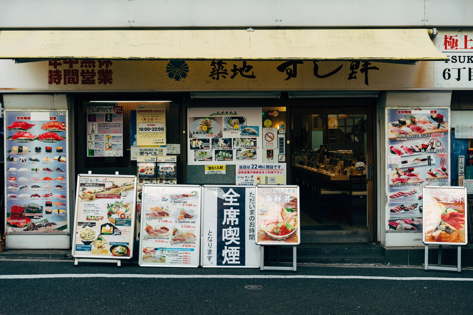The street frontage of a sushi restaurant at Tsukiji, Tokyo, under a yellow awning, with photo menu boards and signs on the sidewalk and the door open to the counter inside.