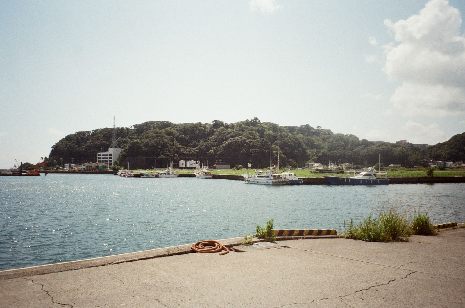 A calm harbor with small boats moored below a wooded headland, a concrete quay in the foreground.