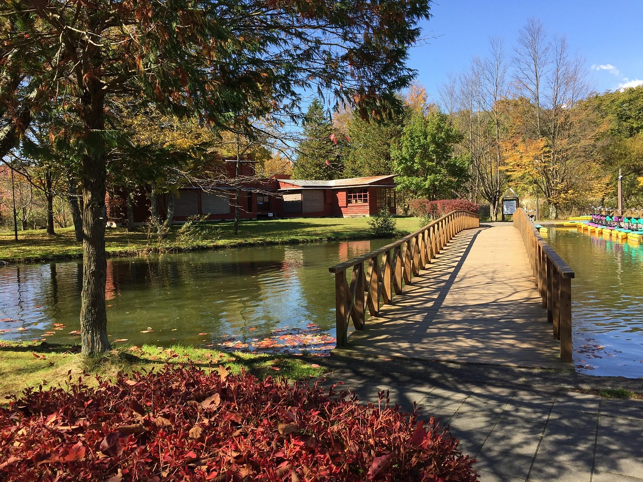 A wooden footbridge across a pond among autumn trees, a low red-roofed building on the far bank and pedal boats moored at the edge.
