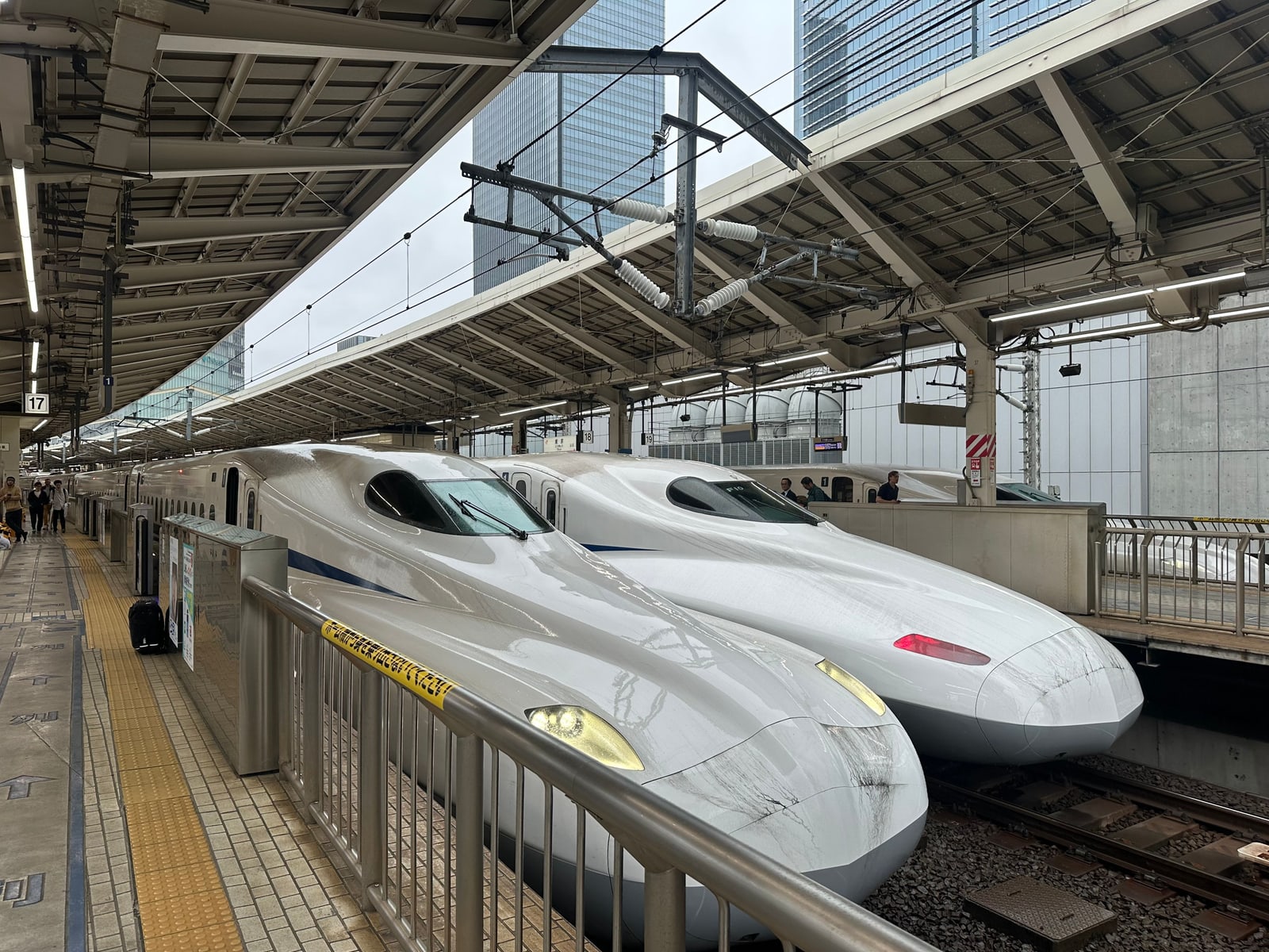 Two white N700-series Shinkansen trains side by side at a covered Tokyo Station platform, high-rise buildings behind.