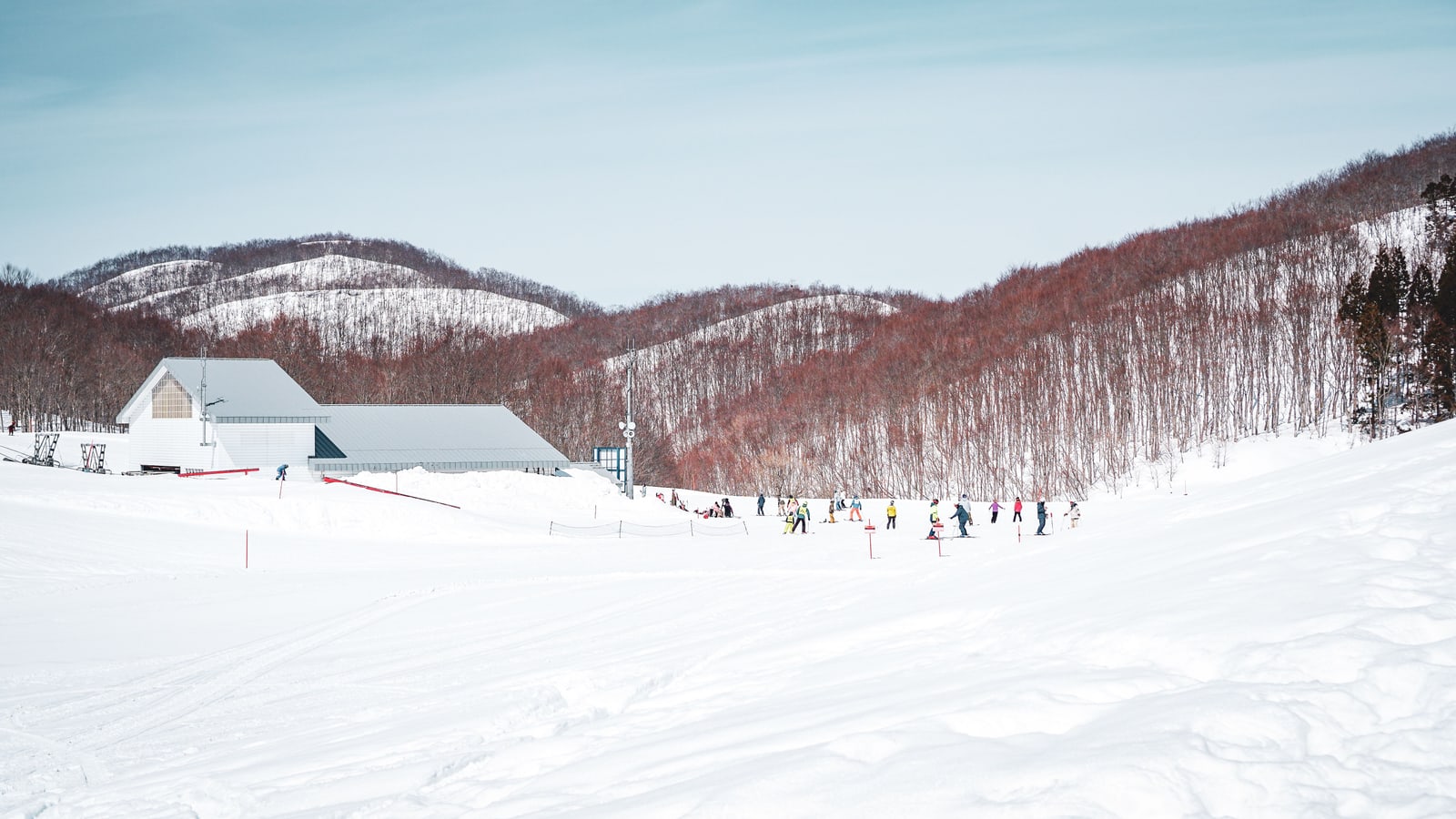 A wide snow-covered ski slope with small distant skiers, a white lodge building and wooded hills behind.