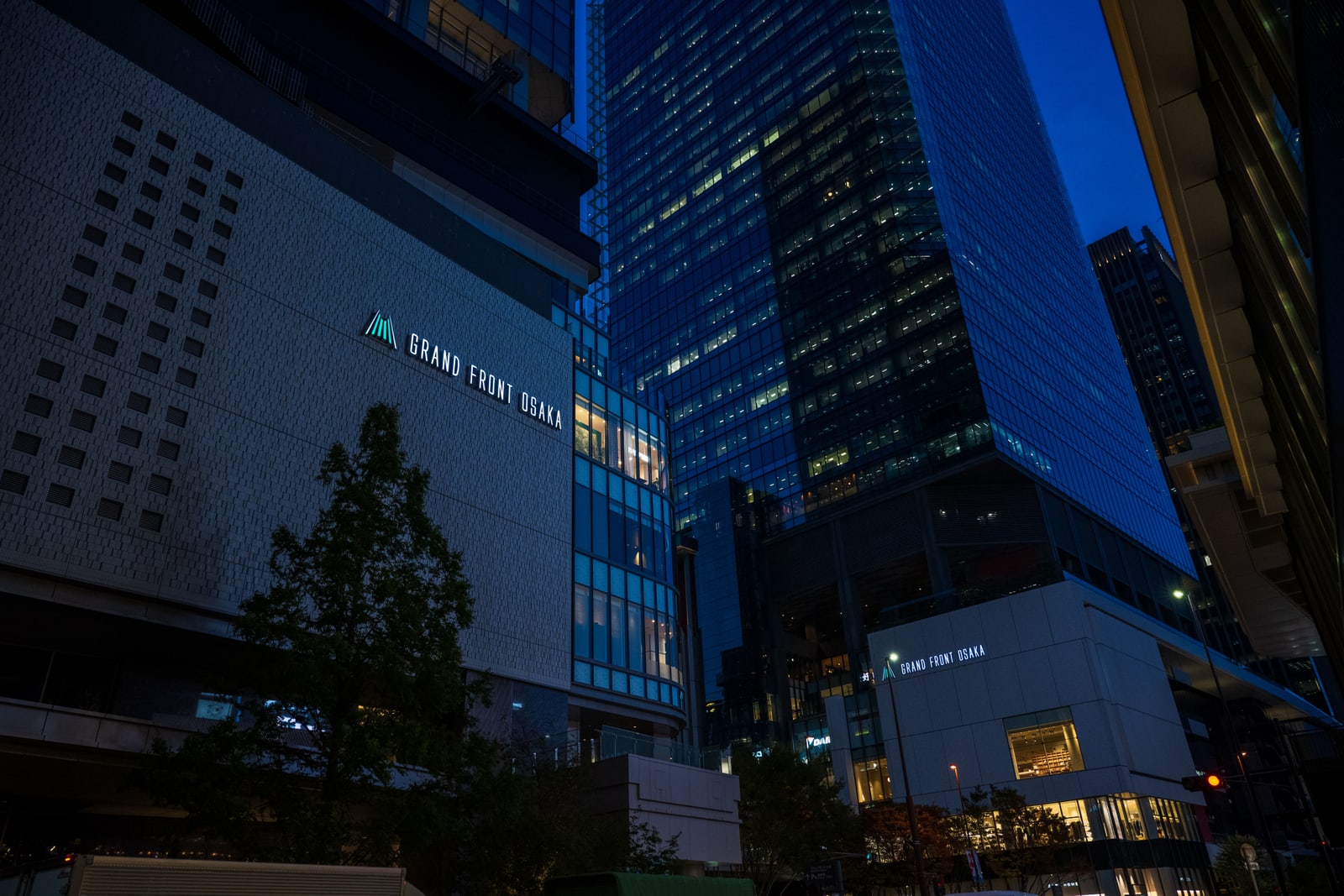 A Grand Front Osaka building at dusk with its lit sign, and a glass office tower rising behind it
