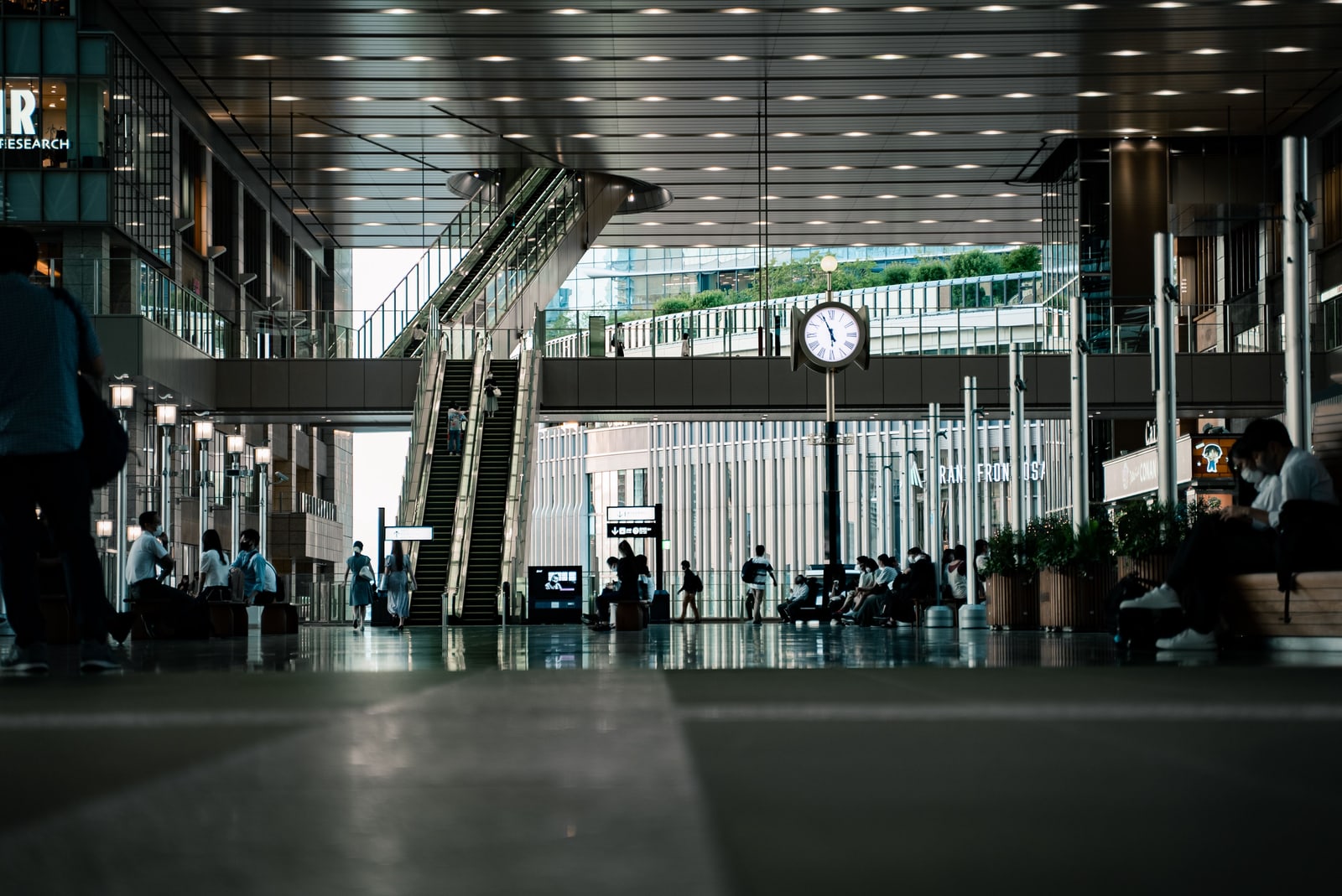 The interior of a train station in Osaka, with escalators, a large clock, and seated commuters.