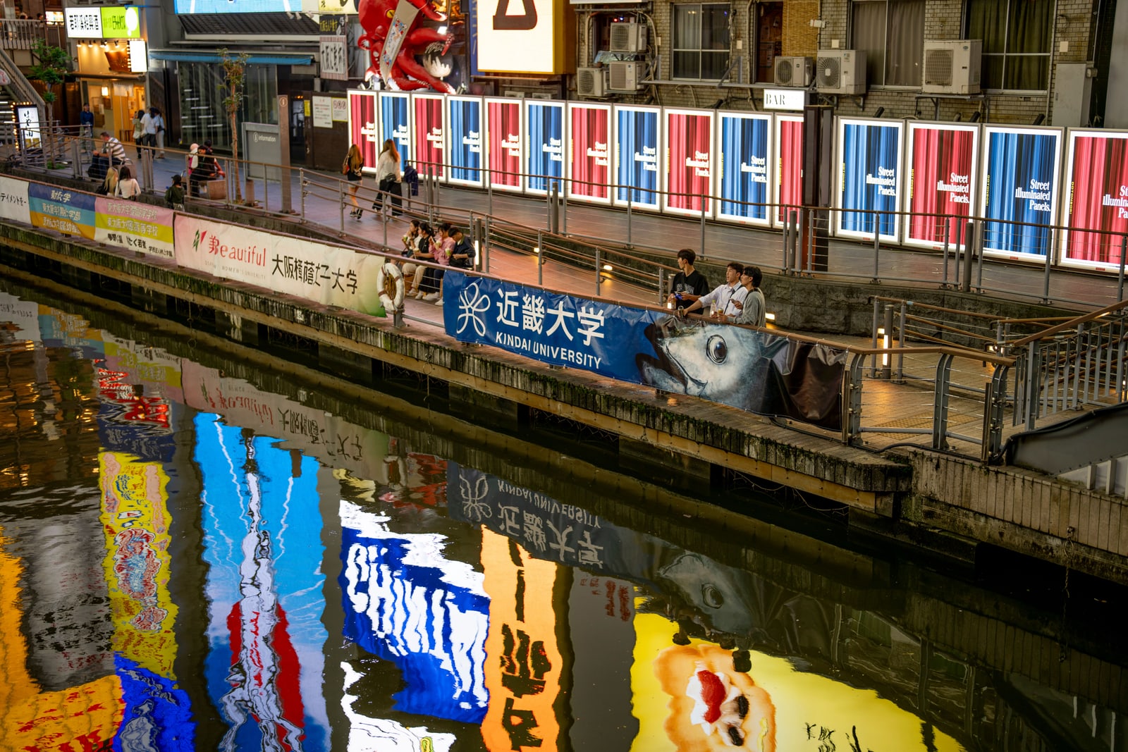 The Dotonbori canal at night, lined with illuminated signs and banners, with pedestrians along the walkway.