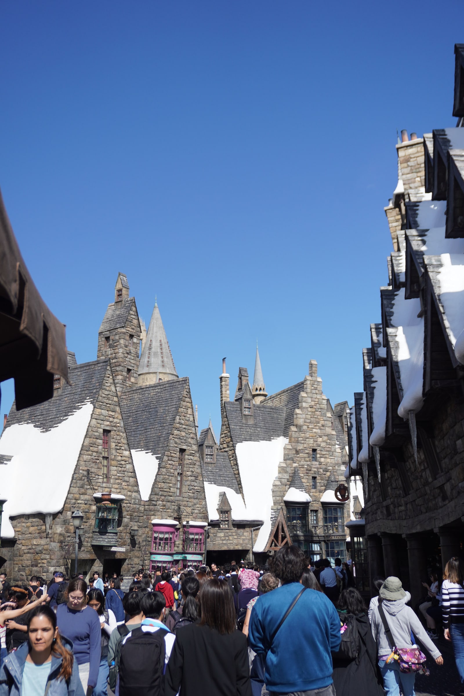 A crowd walks a snow-dusted street between stone buildings at Hogsmeade.
