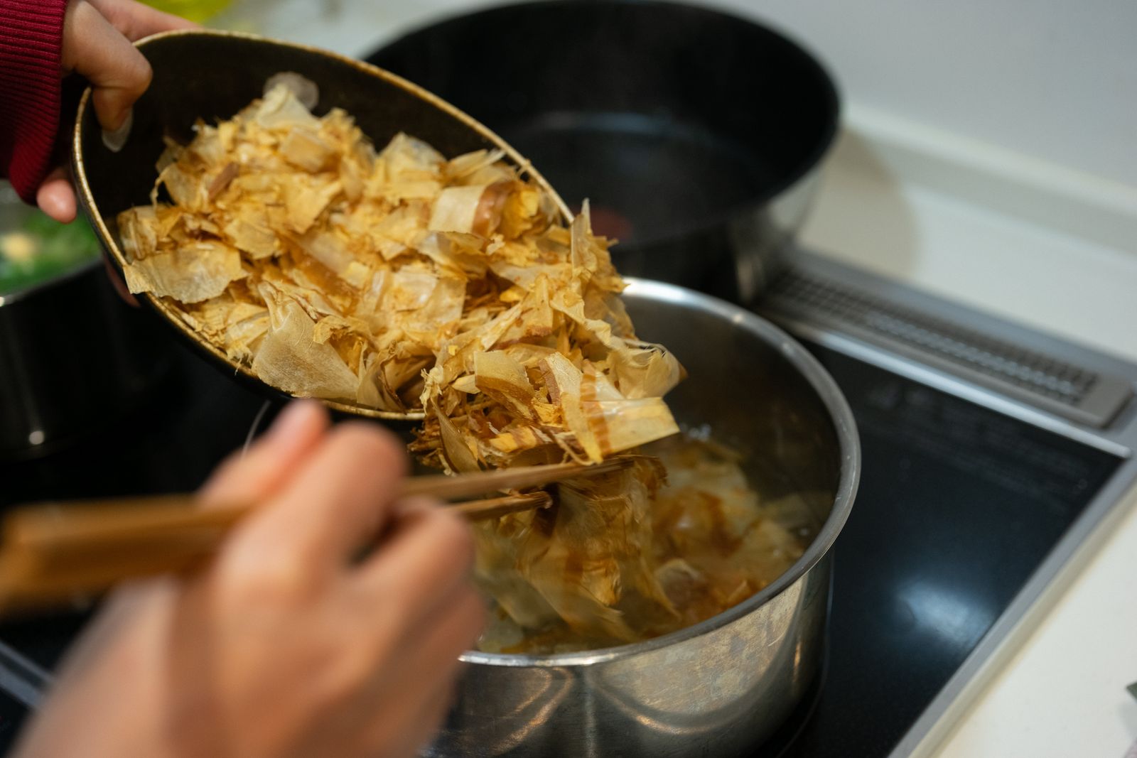Two hands tipping a bowl of dried bonito flakes into a steel pot on a stove, a pair of chopsticks resting in the bowl.