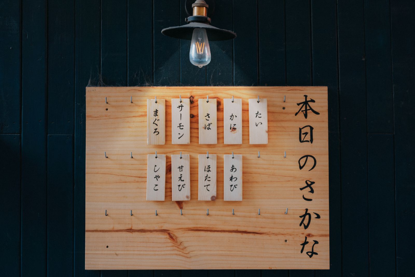 A wooden menu board under a pendant lamp, headed 本日のさかな, with hanging wooden tags naming fish.
