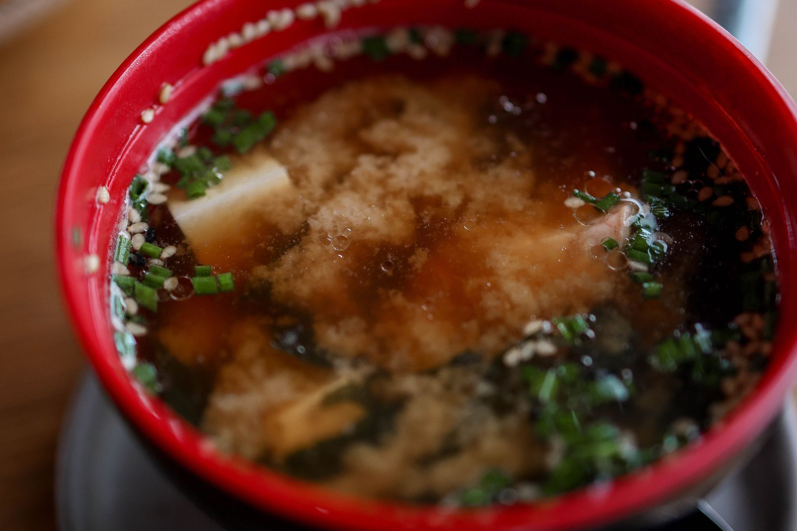 A close view of miso soup in a red lacquer bowl, with cubes of tofu, wakame and chopped chives.