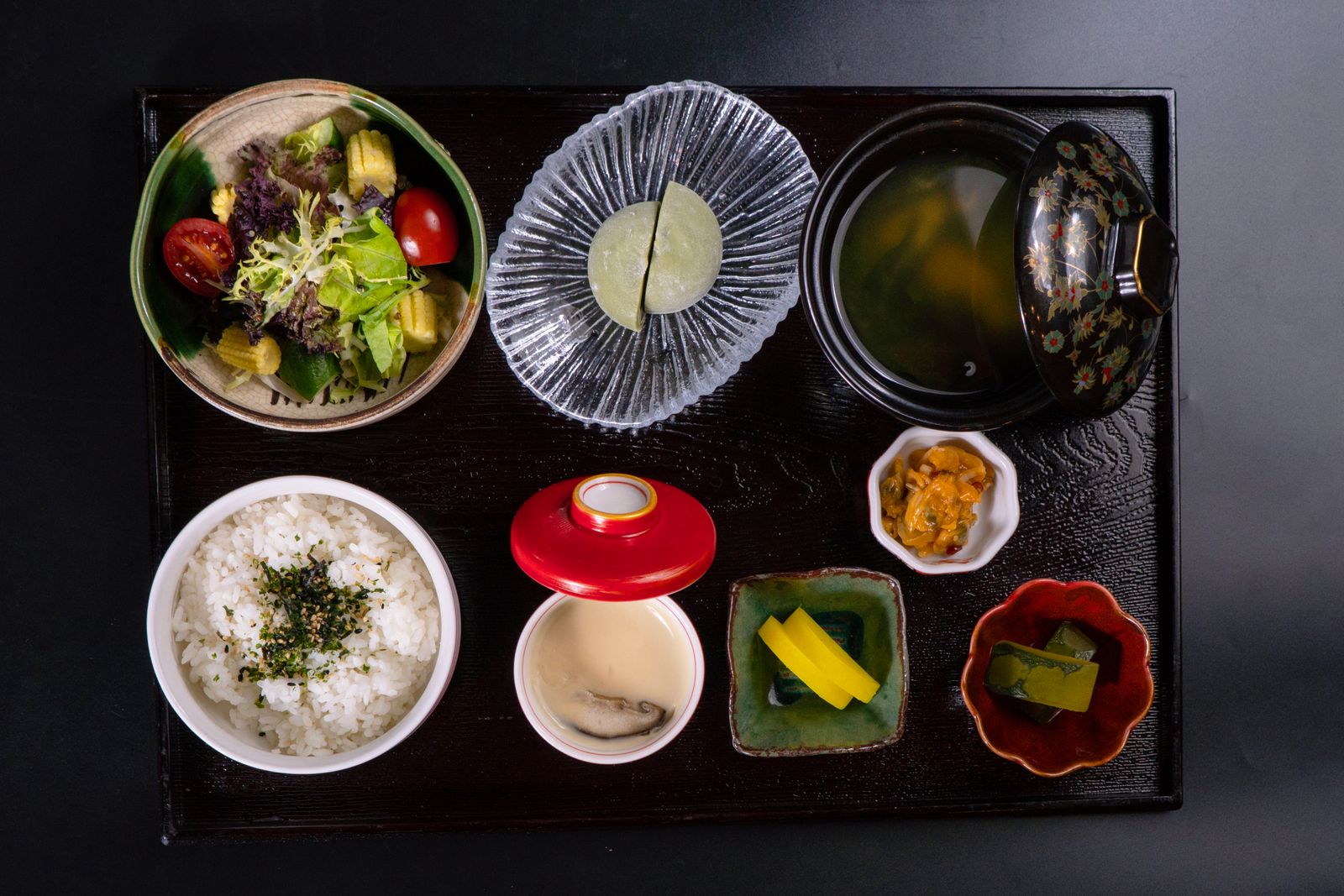 A lacquer tray seen from above: a bowl of rice, a lidded soup bowl, a salad, a cup of chawanmushi, pickles and a small dish of vegetables.