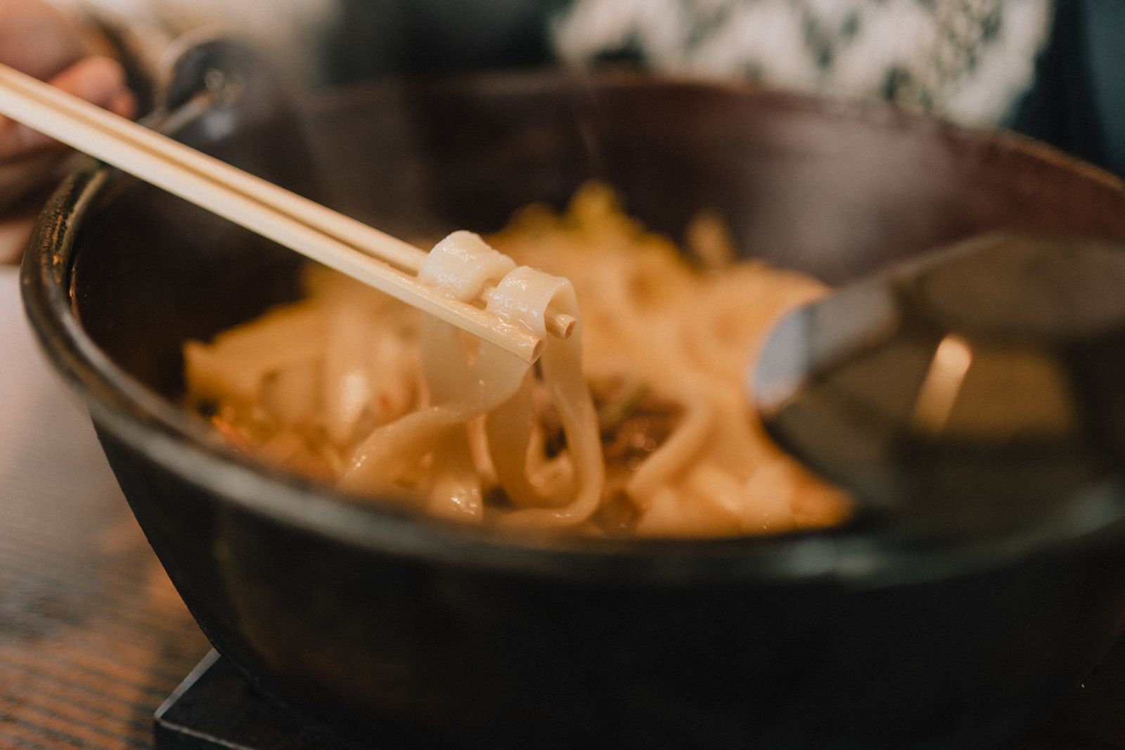 Flat wheat noodles lifted on chopsticks from a hot pot, steam rising, the broth filling the pot below.