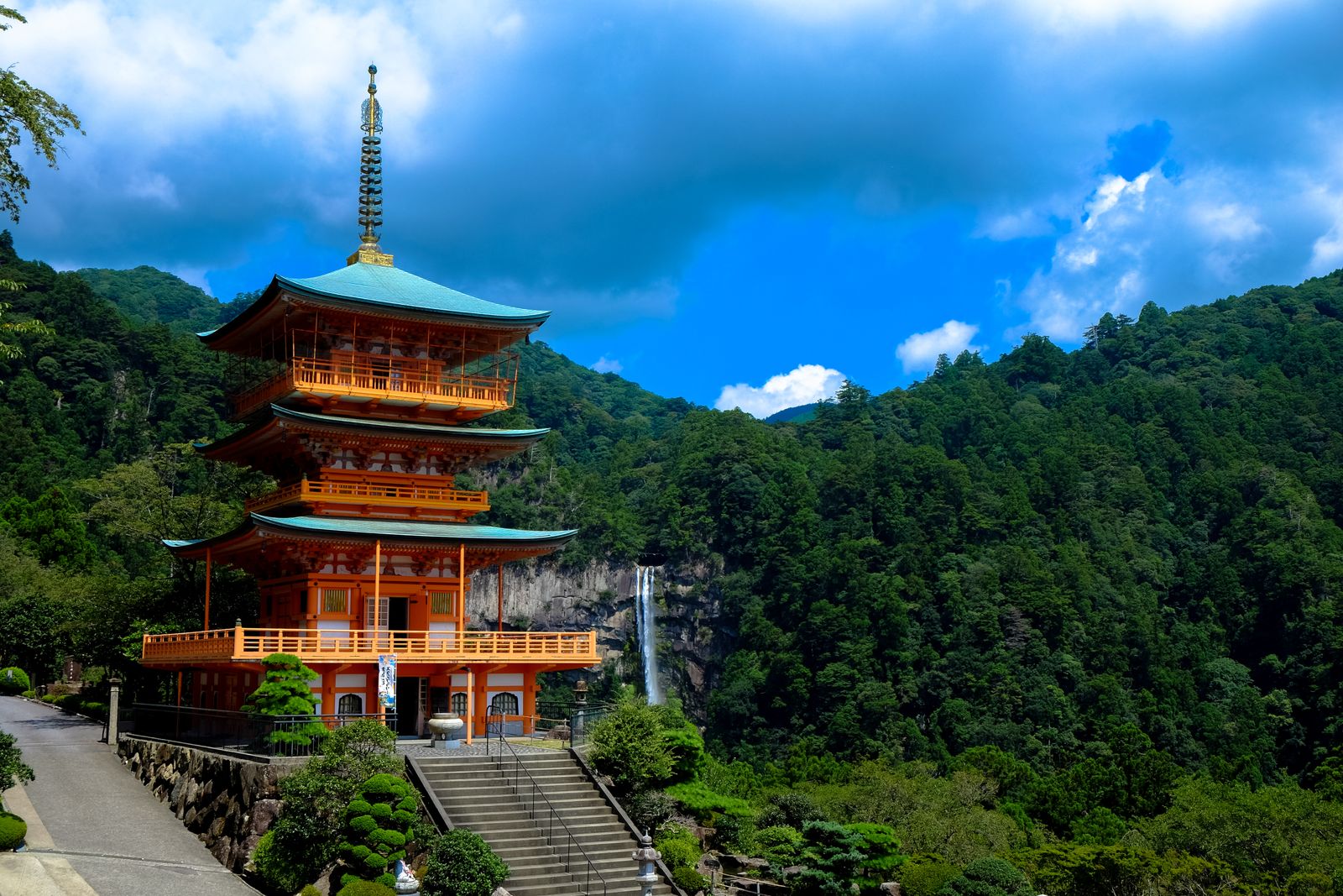 The vermilion three-storey pagoda of Seiganto-ji with Nachi Falls dropping down a grey cliff behind it, forested mountains and blue sky
