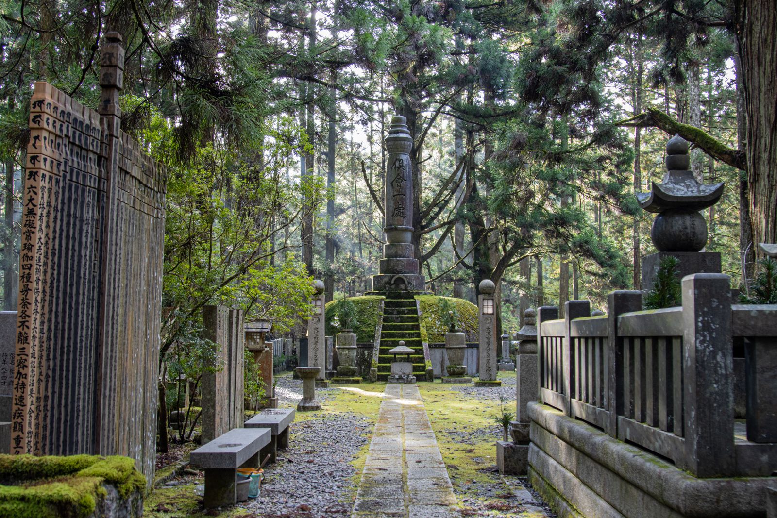 A stone path at Okunoin leading to a tall stone memorial on a mossy mound, wooden grave tablets and lanterns under tall cedars