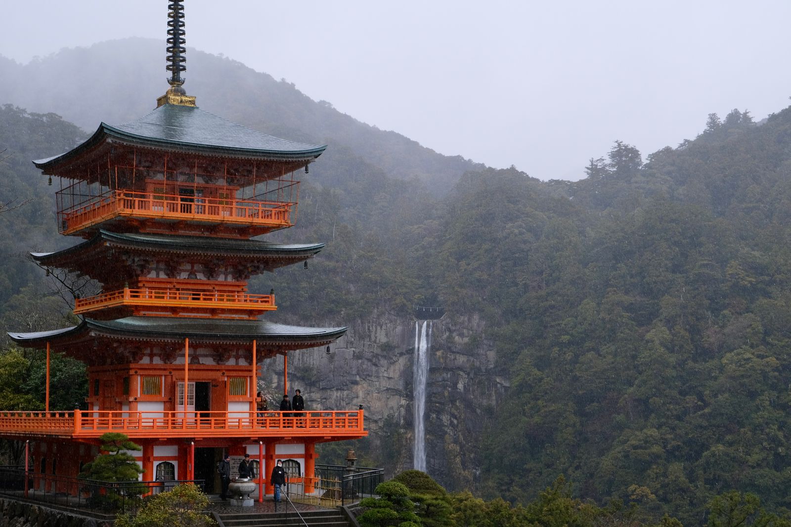 A three-storey vermilion pagoda in rain and mist, a tall waterfall on the cliff behind