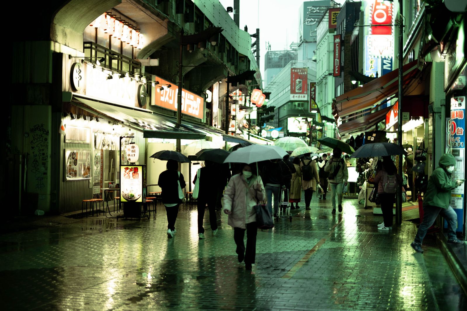 A market street near Ueno Station in the rain, umbrellas up, shops lit, wet pavement reflecting the signs.