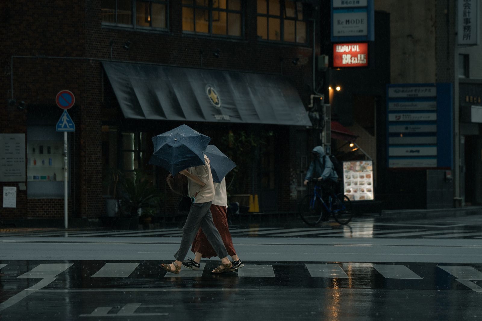 Two people crossing a rain-darkened Tokyo street under umbrellas at dusk, a delivery cyclist waiting behind them.
